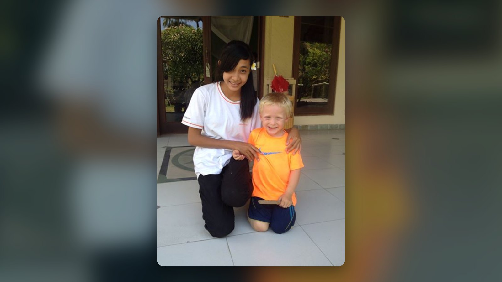 Young girl kneels beside a smiling blond boy in an orange shirt outside a building, with her arm around him.